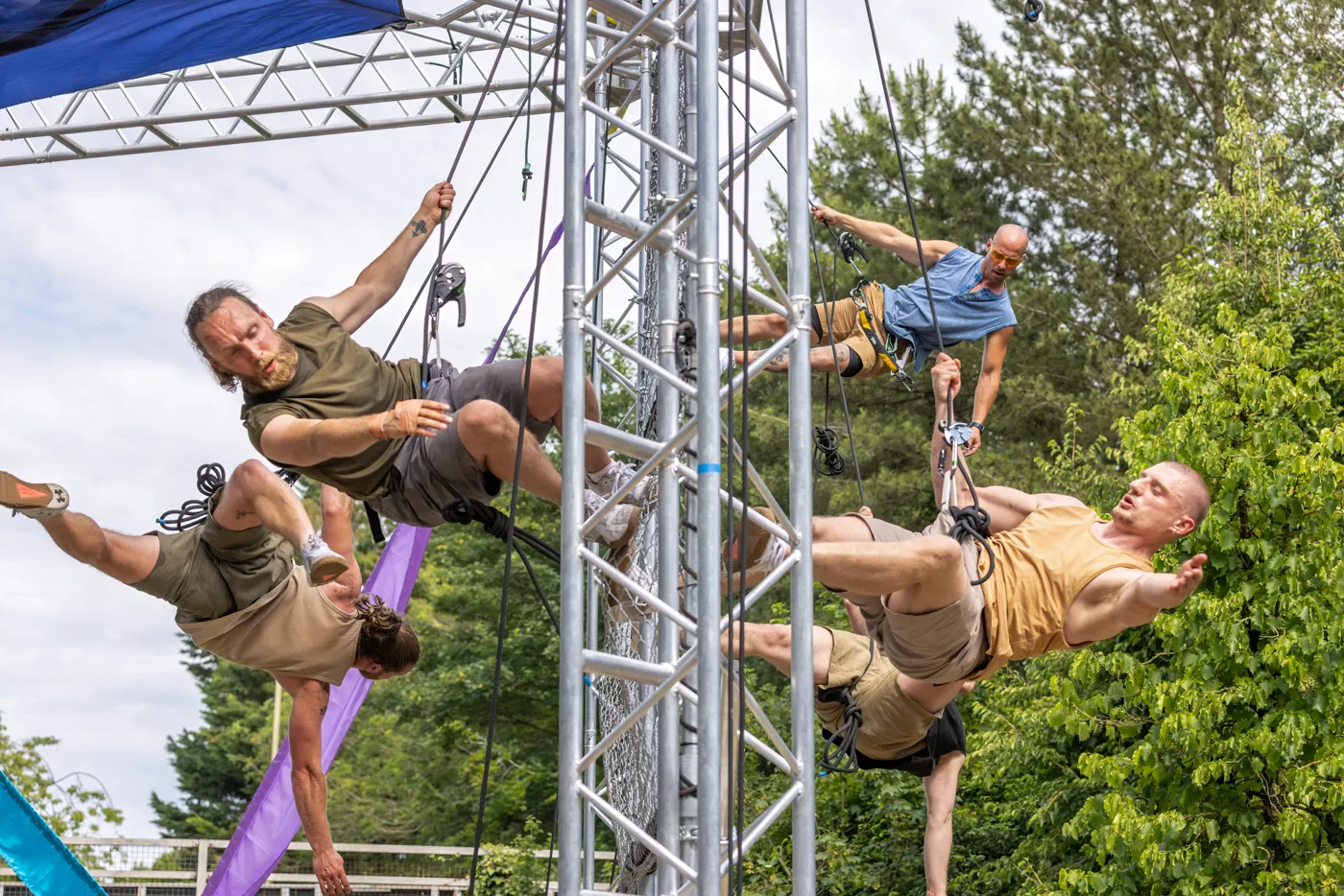 Four Autin Dance Theatre aerialists in harnesses hanging from the metal truss of the Up in the Sky rig, with trees behind
