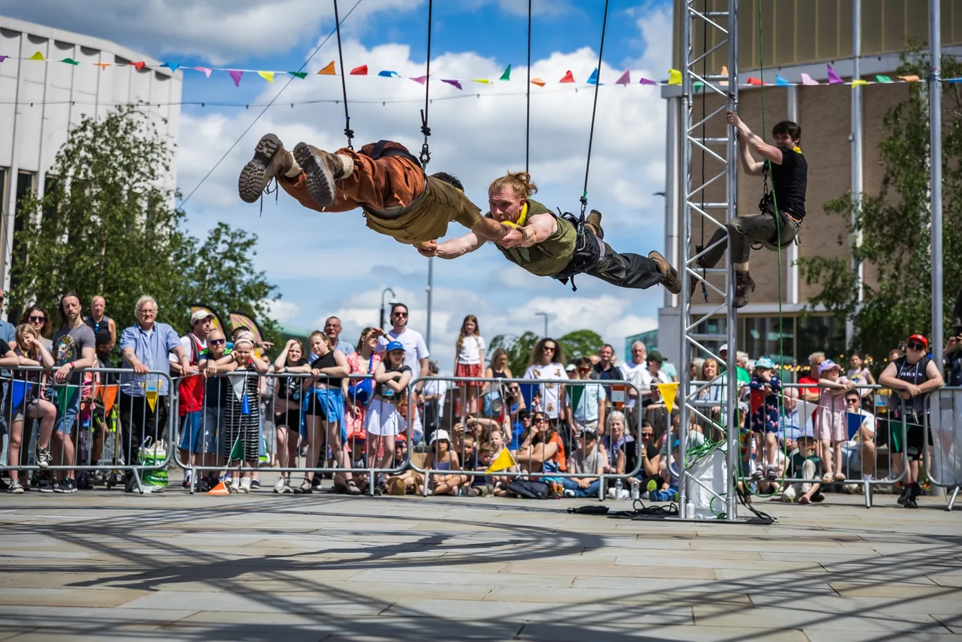 Two aerialists flying horizontally on ropes above a crowd of families behind barriers in a paved square hung with bunting, with a third performer climbing a metal tower