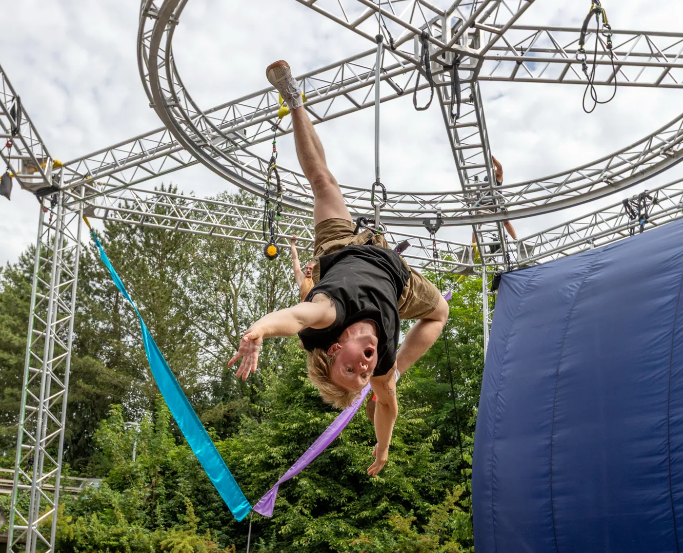 A performer in a harness swinging from a rope beside a blue backdrop and green trees, one hand on a purple fabric ribbon